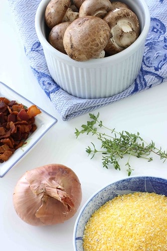 Bowls of mushrooms, polenta and bacon, and an onion, on a white surface.