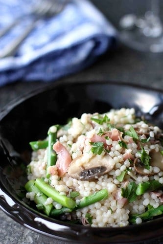 Israeli couscous in a black bowl with asparagus, prosciutto and cooked mushrooms.