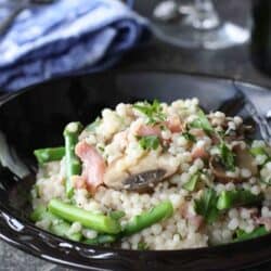 Pearl couscous, asparagus, prosciutto and mushrooms in a black bowl.