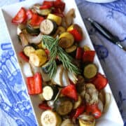 Balsamic roasted vegetables and rosemary on a white platter and blue tablecloth.