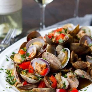 Steamed clams, sausage and red pepper in a white bowl, with glasses of wine in background.