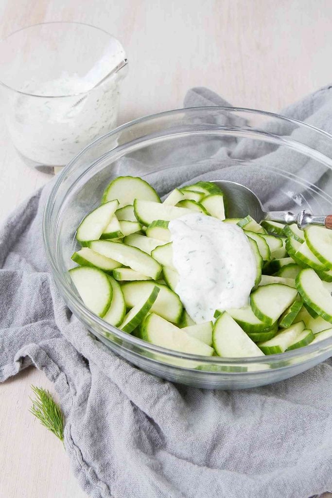 Sliced cucumber in a glass bowl, with dill yogurt dressing poured over top.