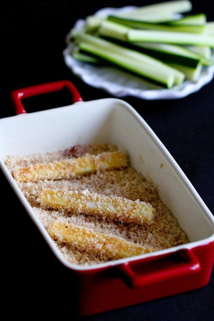 Zucchini sticks in a container of panko breadcrumbs.
