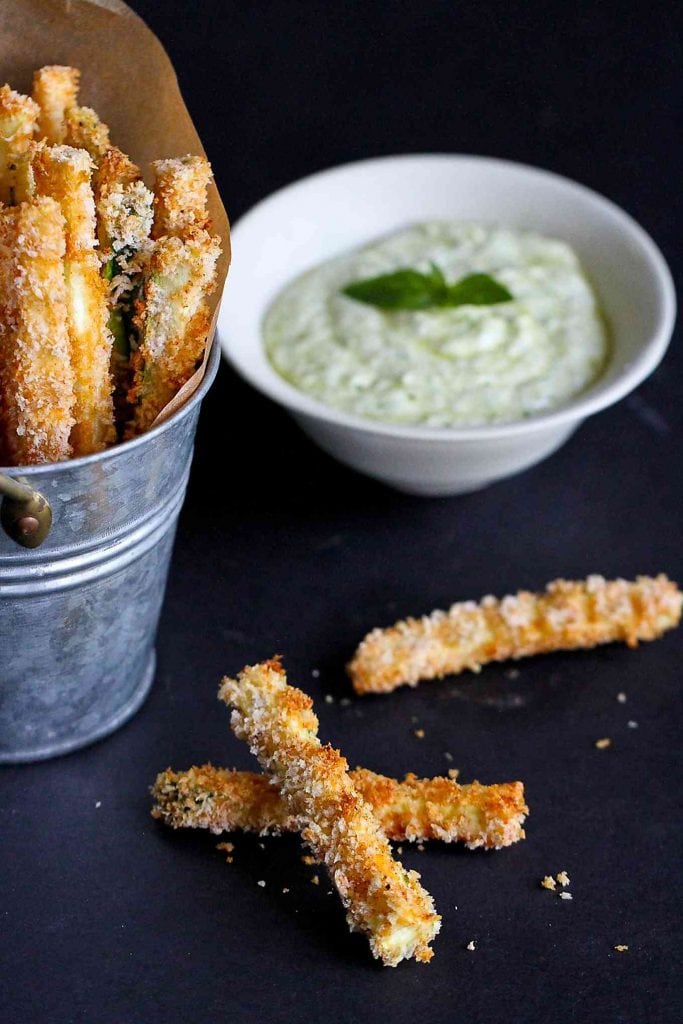 Baked zucchini fries in a metal container and on a dark surface, plus dish of yogurt dip.