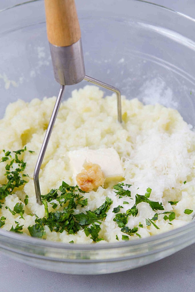 Mashed cauliflower, roasted garlic, butter and parsley in a glass bowl.