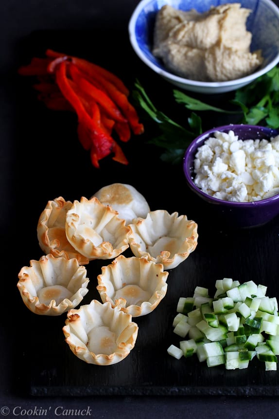 Phyllo cups, feta cheese, cucumber, hummus and roasted peppers on a cutting board.