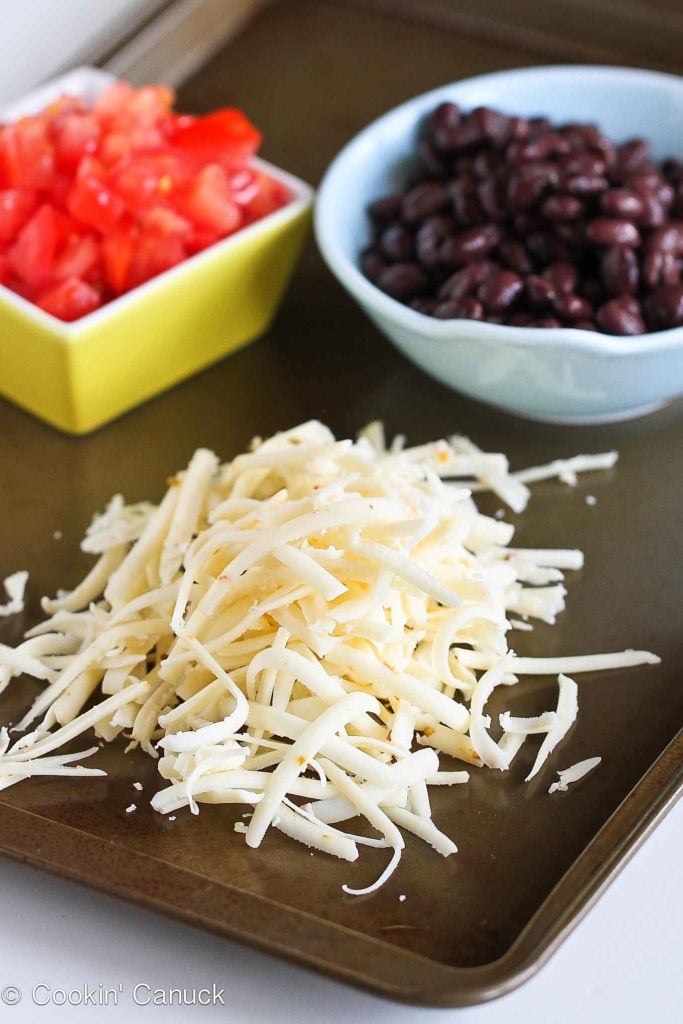 Shredded pepper Jack cheese on a cutting board. Black beans and chopped tomatoes in bowls.