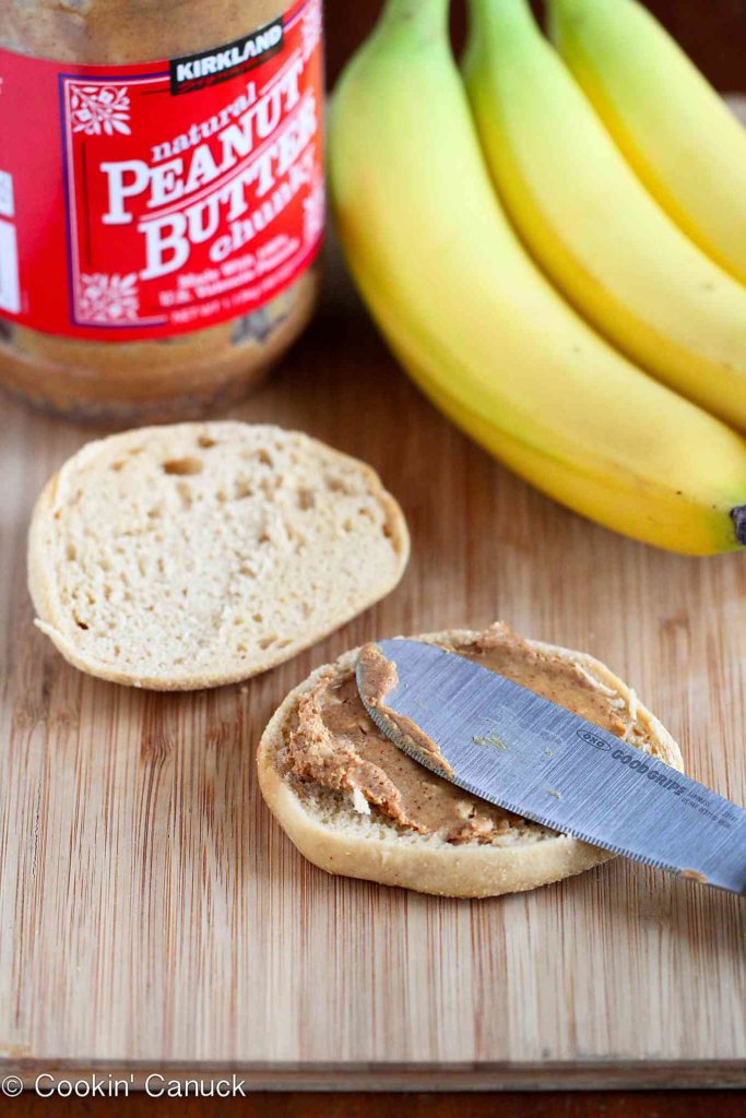 Spreading peanut butter on an English muffin, plus bananas, all on a wooden cutting board.