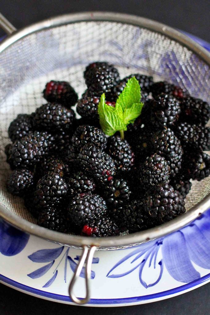Blackberries in a fine mesh sieve, sitting over a blue plate.