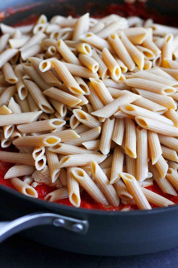 Cooked whole wheat penne sitting on a tomato sauce in a large nonstick skillet.