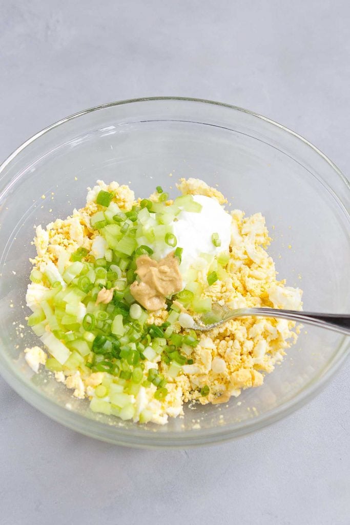 Chopped hard boiled egg, mayonnaise, celery, green onion and Dijon mustard in a glass bowl.