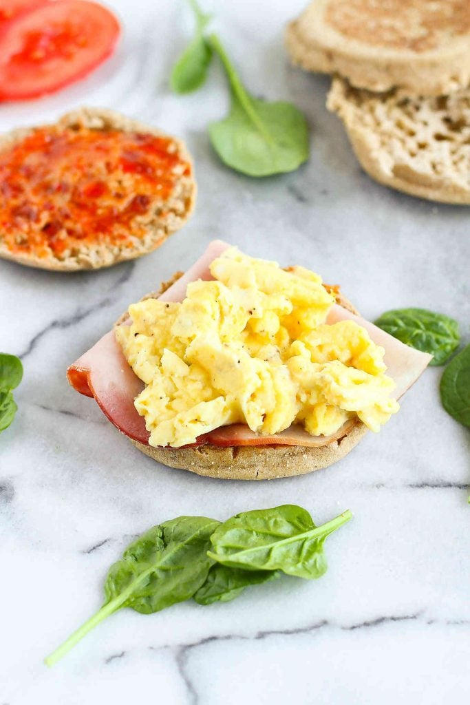 Scrambled eggs on top of an open English muffin. Sitting on a marble slab, with spinach leaves.