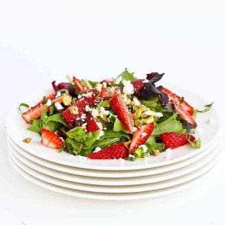 Green salad with strawberries, feta cheese and pistachios on a stack of white plates.
