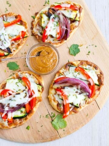Overhead photo of Thai veggie naan bread pizzas on a pizza peel, with a bowl of peanut sauce.