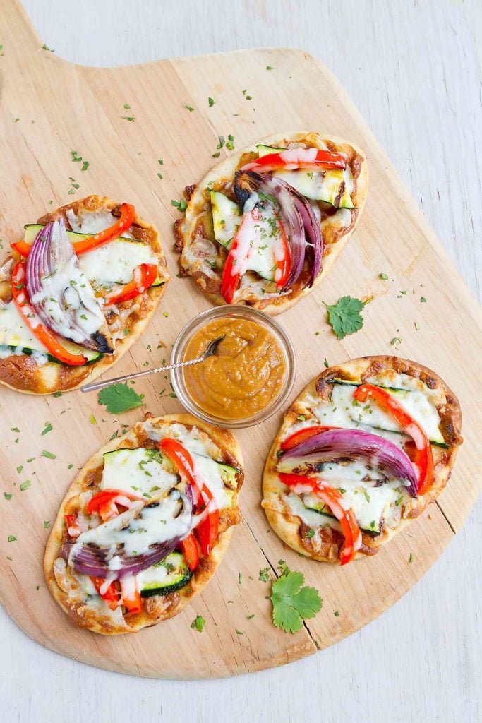 Thai vegetarian naan pizzas topped with grilled vegetables, peanut sauce and melted cheese, on a wooden pizza peel.