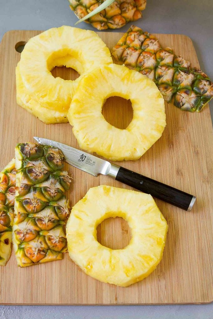 Slices of fresh pineapple on a bamboo cutting board. Top of pineapple in background.