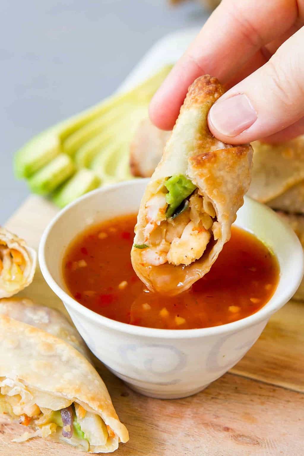 Half of a shrimp egg roll being dipped into a bowl of sweet chili sauce. Slices of avocado behind.