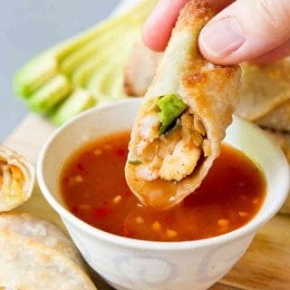 Half of a shrimp egg roll being dipped into a bowl of sweet chili sauce. Slices of avocado behind.