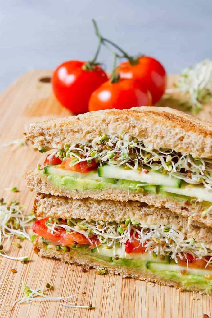 Two stacked sandwich halves, filled with avocado, cucumber, tomato and sprouts. Tomatoes in the background.