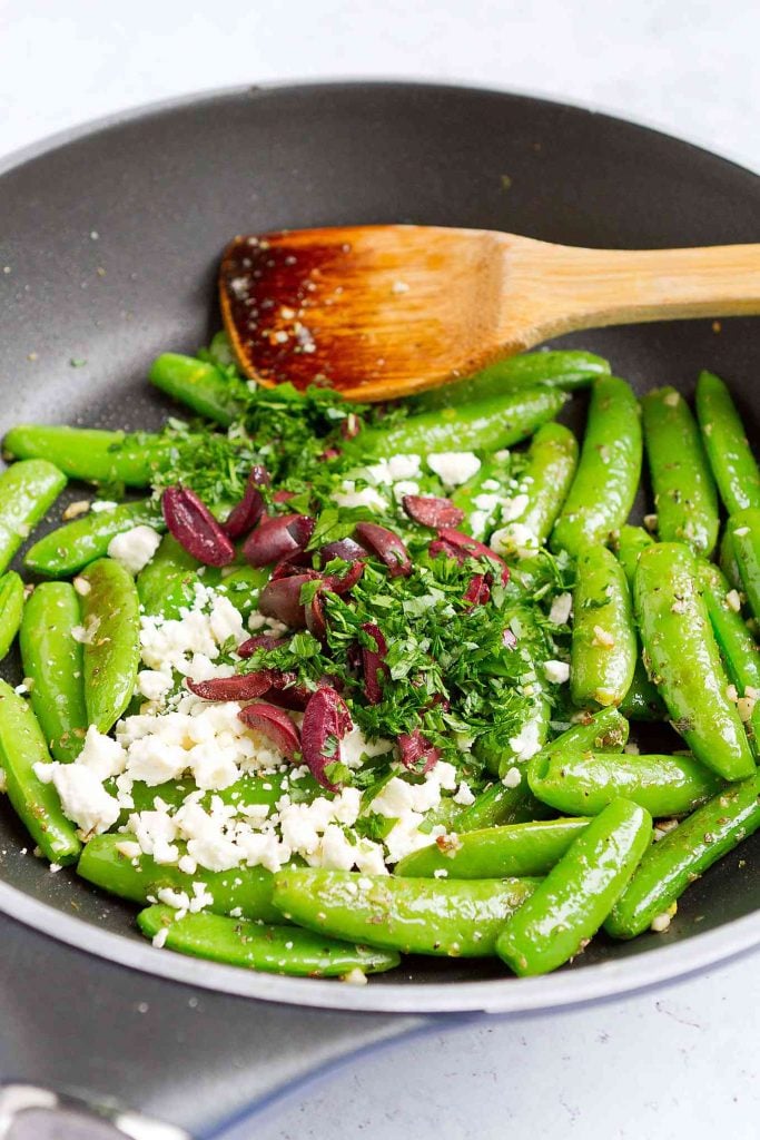 Sugar snap peas, feta and kalamata olives in a nonstick skillet with a wooden spoon.