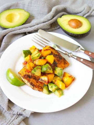 Grilled fish with avocado peach salsa on a white plate, plus two half avocados in background.