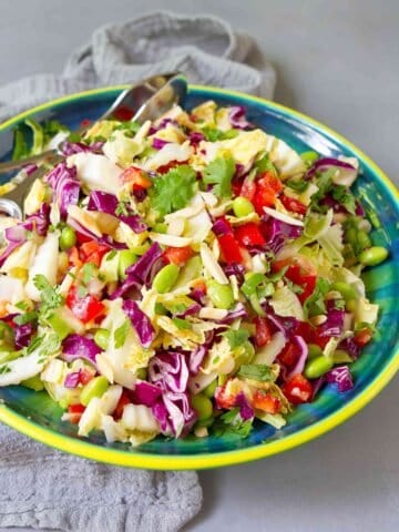 Slaw with green and red cabbage, bell pepper, edamame, cilantro and almonds in a blue-green bowl, on a gray napkin.