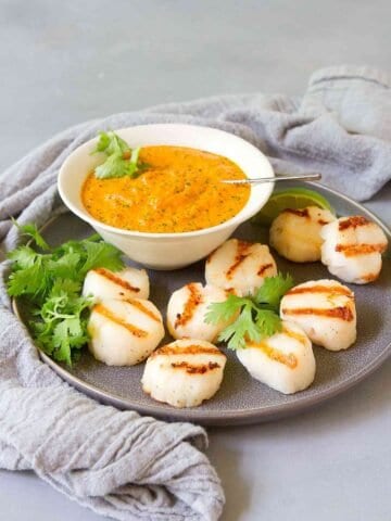 Grilled scallops on a gray plate with a bowl of red pepper pesto. Plus, cilantro leaves.