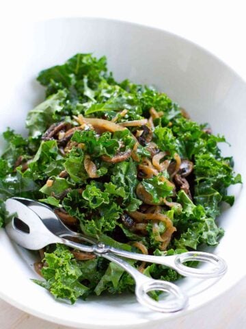 Kale salad with caramelized onions and mushrooms in a white bowl with silver tongs.