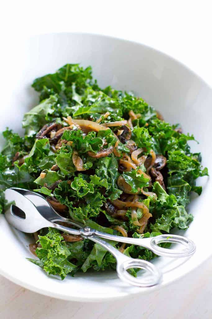 Kale salad with caramelized onions and mushrooms in a white bowl with silver tongs.
