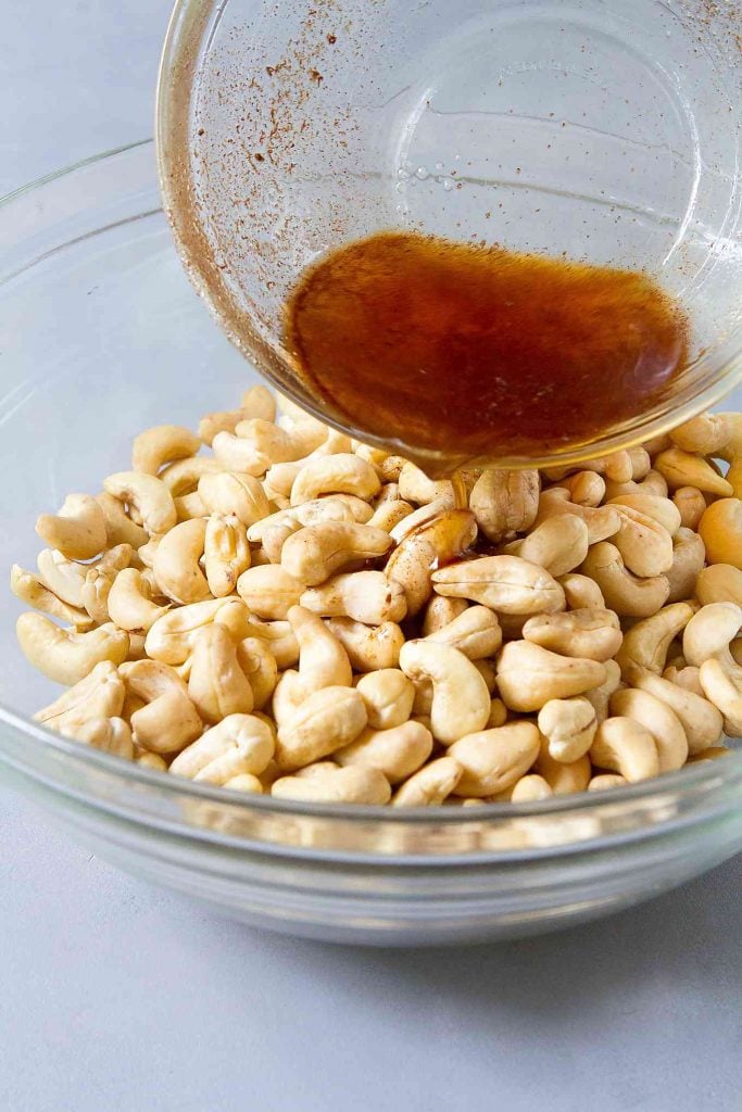 Cashews in a glass bowl, pouring maple syrup & cinnamon mixture over top.