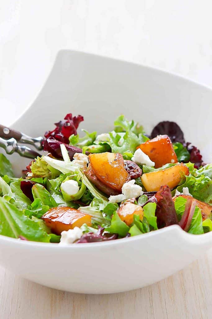 Green salad in a large white bowl, with golden brown pears, goat cheese and wooden salad servers.