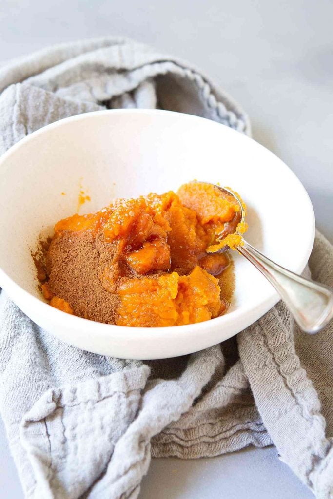 Pumpkin puree in a white bowl, with maple syrup, ground cinnamon and a spoon. Gray napkin on side.