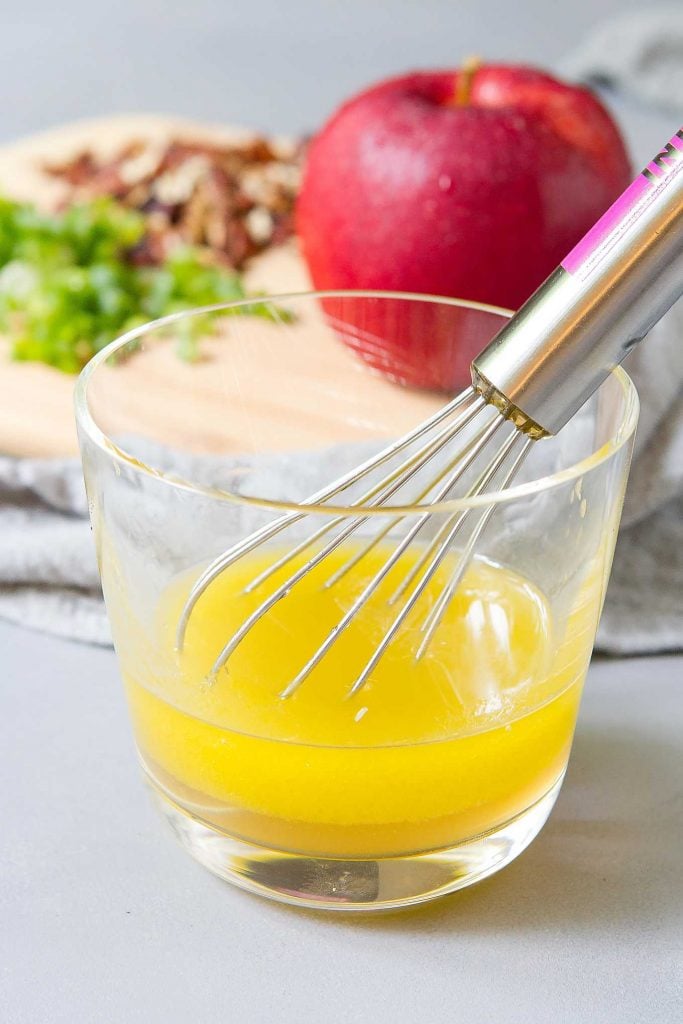 Apple cider vinaigrette in a glass with a whisk. Apple and pecans in the background.