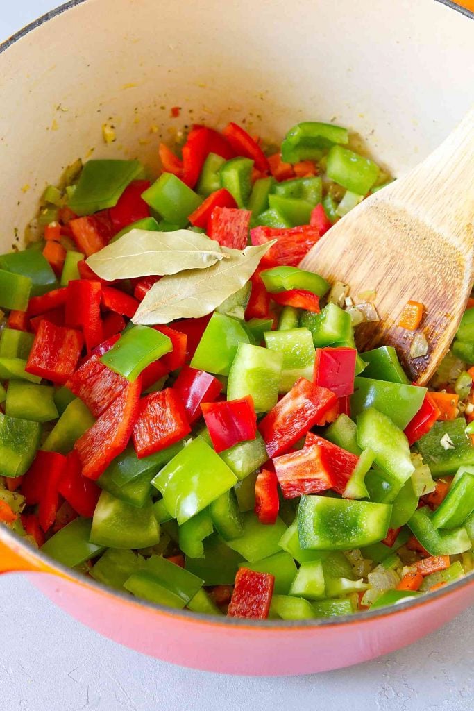 Chopped red and green bell peppers, bay leaves and metal spatula in a large orange saucepan.