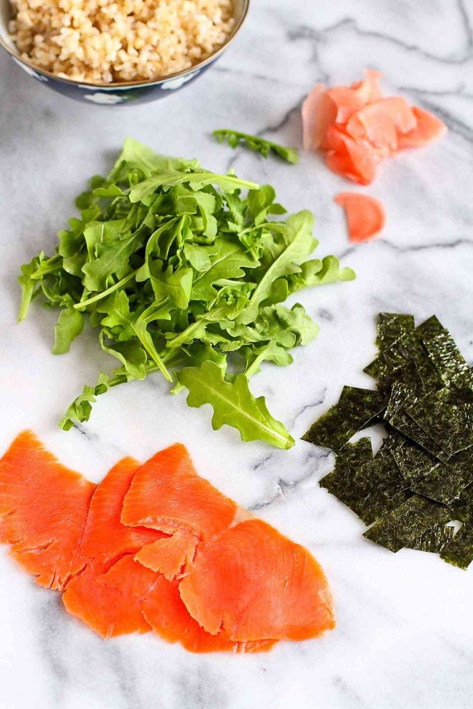 Arugula, smoked salmon, nori sheets, pickled ginger and rice on a marble surface.