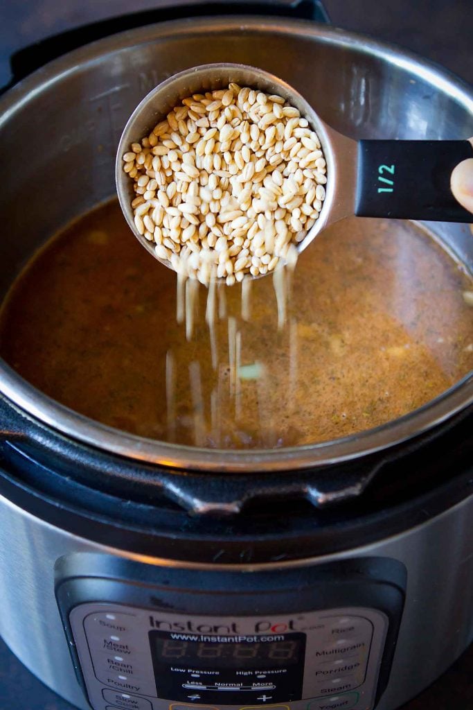 Pouring dry barley into an Instant Pot filled with broth, beef and vegetables.