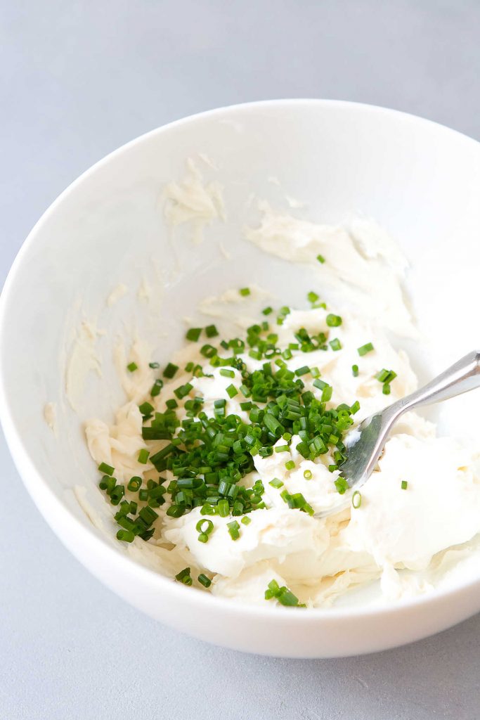 Cream cheese with chopped chives and a stainless steel spoon in a white bowl