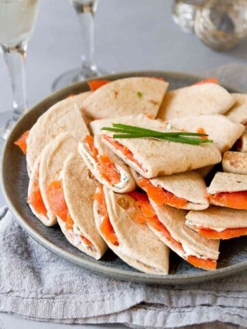 Mini quesadillas filled with cream cheese and smoked salmon, piled on dark gray plate. Glass of Prosecco behind.