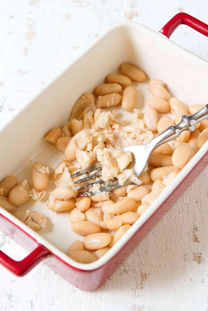 Small red ceramic dish with partially smashed white beans, with a silver fork.