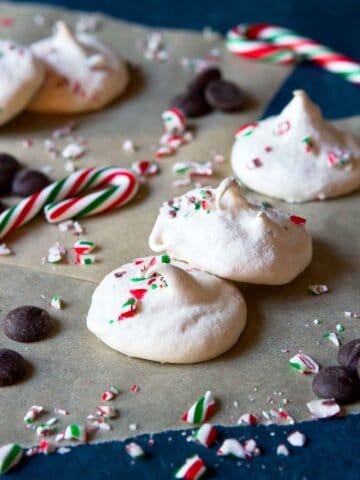 Several meringue cookies on parchment paper, surrounded by some mini candy canes and chocolate chips