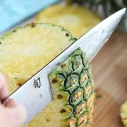 A large chef's knife slicing the skin from a pineapple.