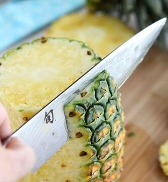 A large chef's knife slicing the skin from a pineapple.
