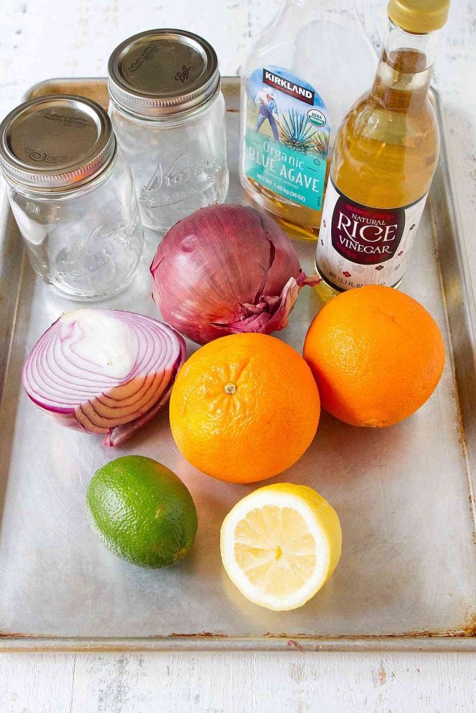 Red onions, oranges, lime, lemon, vinegar and jars on a baking sheet.