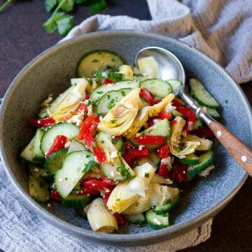 Cucumber, roasted red peppers and artichokes in a gray bowl, with parsley in background.