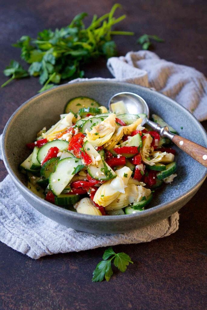 Cucumber, roasted red peppers and artichokes in a gray bowl, with parsley in background.