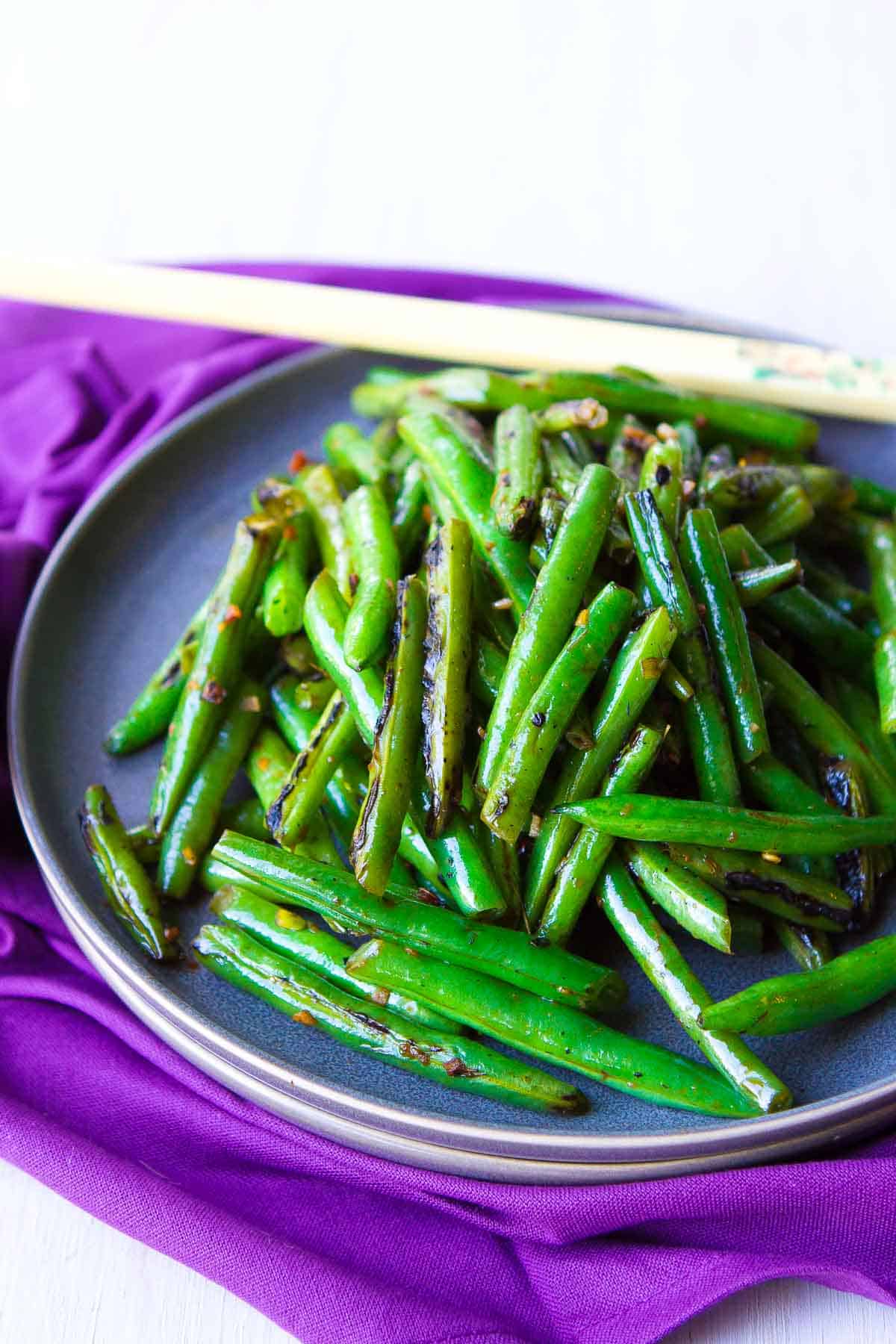 Cooked green beans and chopsticks on a dark gray plate, resting on a purple napkin.