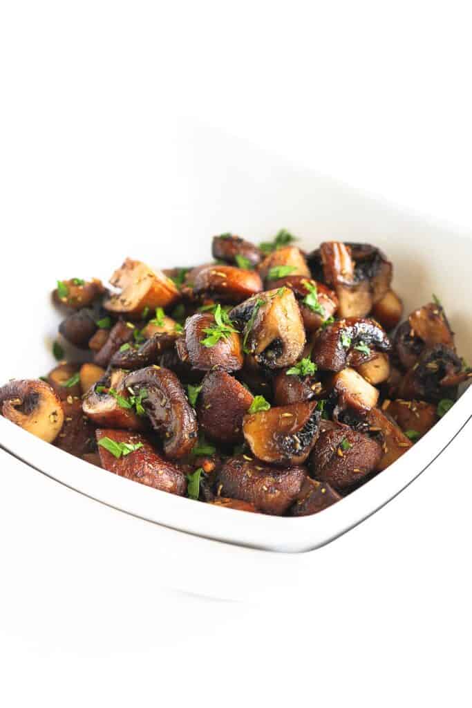 Roasted mushrooms, cut in quarters, in white bowl. Topped with parsley.