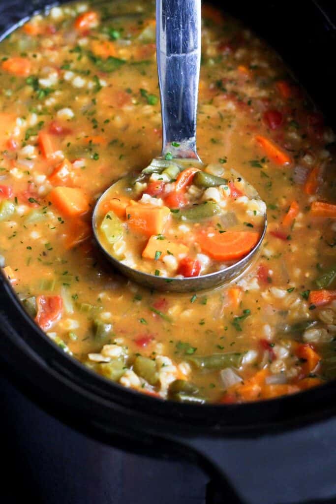 A ladle scooping vegetable barley soup out of a crockpot.