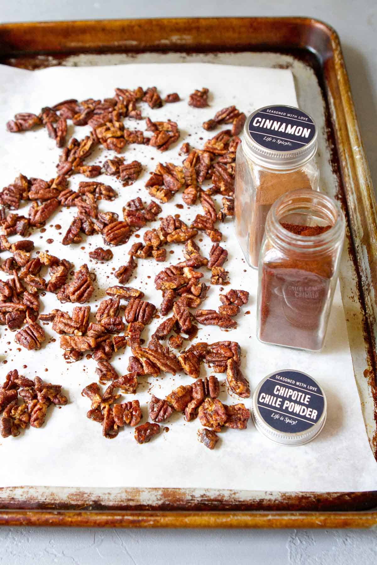 Chopped spiced pecans on a baking sheet lined with parchment paper. Spice bottles on the side.