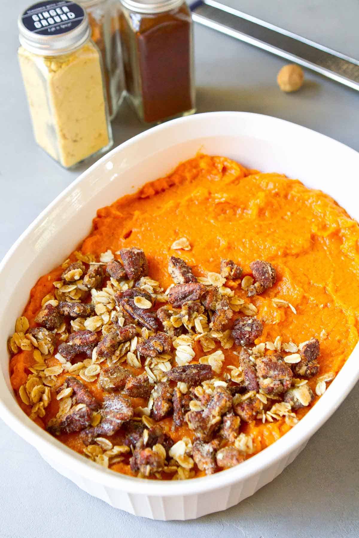 Mashed sweet potatoes in a white baking dish, partially covered with a pecan streusel topping. Spice bottles on the side.